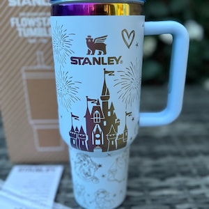 May include: White Stanley Flowstate tumbler with a blue handle and lid. The tumbler features a castle design with fireworks and the Stanley logo. A heart and other graphics are also visible.