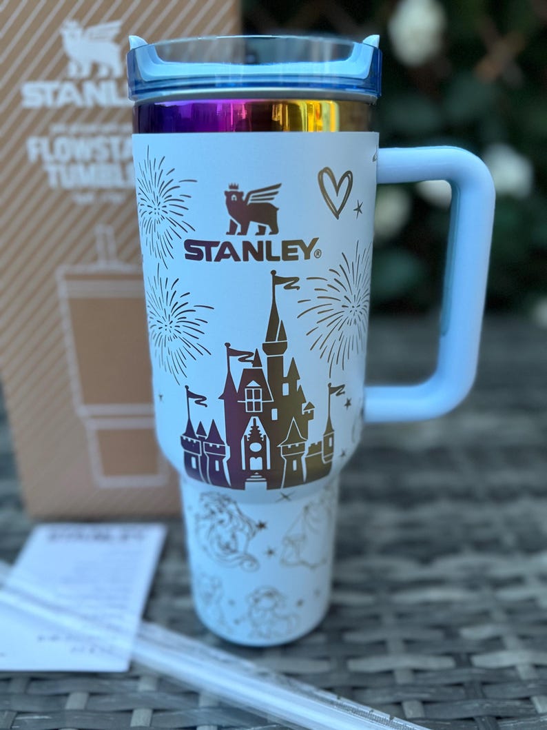 May include: White Stanley Flowstate tumbler with a blue handle and lid. The tumbler features a castle design with fireworks and the Stanley logo. A heart and other graphics are also visible.