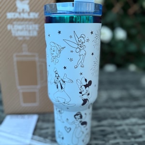 May include: A light blue Stanley Flowstate tumbler with a clear straw. The tumbler features black and white illustrations of Disney characters, including Peter Pan, Tinkerbell, Goofy, Minnie Mouse, and a princess. The lid is blue.