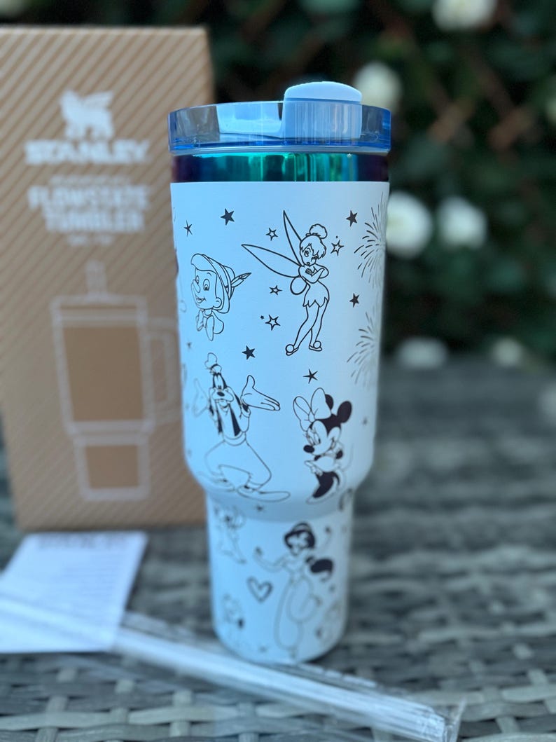 May include: A light blue Stanley Flowstate tumbler with a clear straw. The tumbler features black and white illustrations of Disney characters, including Peter Pan, Tinkerbell, Goofy, Minnie Mouse, and a princess. The lid is blue.