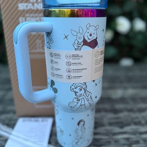 May include: A light blue insulated tumbler with a handle, featuring illustrations of Disney characters. The tumbler has a blue lid and a rainbow-colored band. A paper tag with product information is wrapped around the tumbler.
