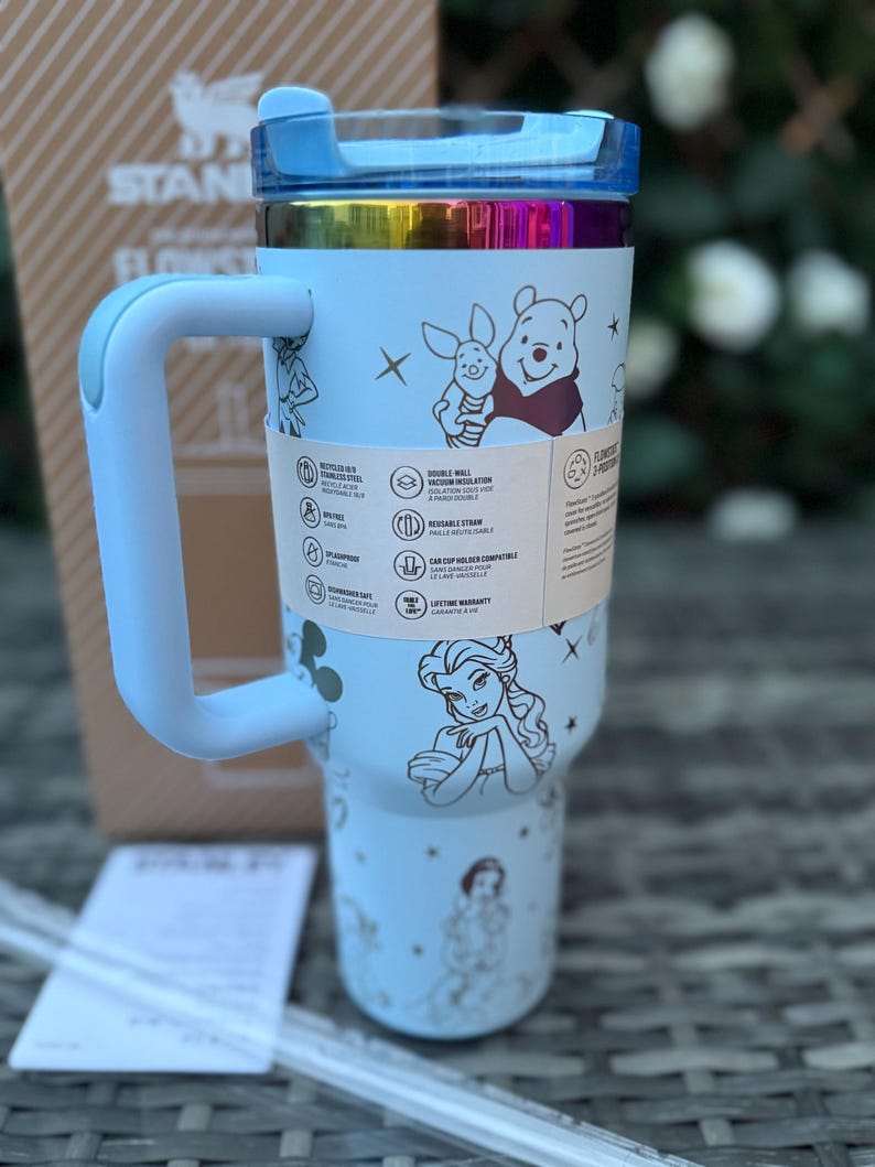 May include: A light blue insulated tumbler with a handle, featuring illustrations of Disney characters. The tumbler has a blue lid and a rainbow-colored band. A paper tag with product information is wrapped around the tumbler.