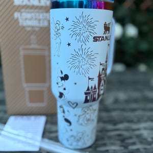 May include: A light blue Stanley Flowstate tumbler with a clear straw. The tumbler features illustrations of Tinkerbell, Mickey Mouse, fireworks, a castle, and other Disney-themed designs. The lid is blue and iridescent.