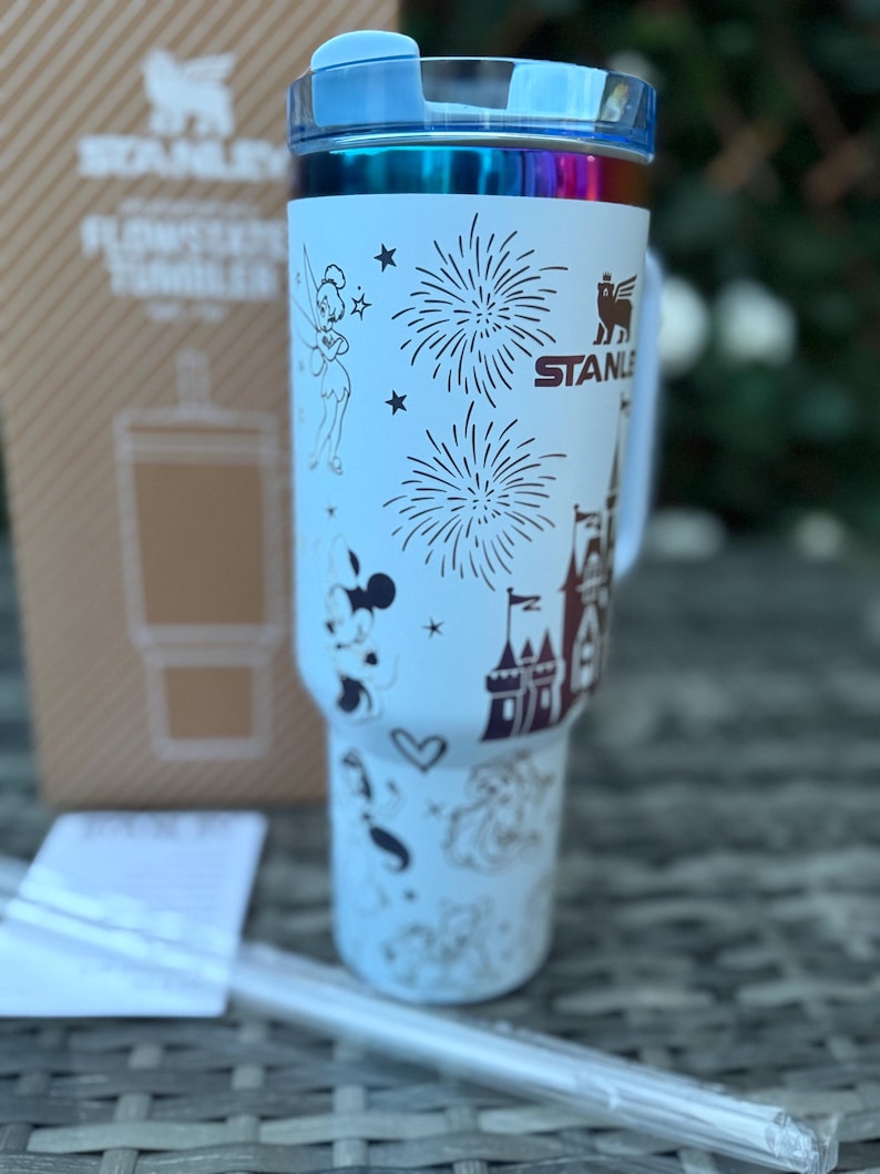 May include: A light blue Stanley Flowstate tumbler with a clear straw. The tumbler features illustrations of Tinkerbell, Mickey Mouse, fireworks, a castle, and other Disney-themed designs. The lid is blue and iridescent.