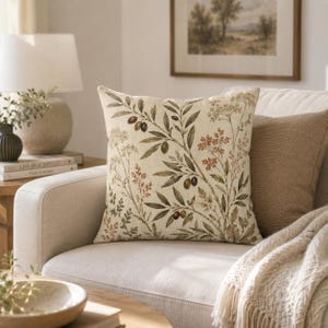 May include: A square throw pillow featuring a botanical print on a cream-colored background. The design displays olive branches with dark green and brown olives, interspersed with delicate sprigs of dried wildflowers and foliage in earthy tones.