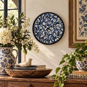 May include: A round wall clock with a black frame and a dark blue face featuring a white floral pattern. The clock has black hour, minute, and second hands, and black Arabic numerals from one to twelve positioned around the circular dial.