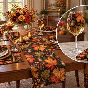 May include: A clear glass wine goblet featuring a decorative wrap around the upper bowl with a black background, orange and yellow autumn maple leaves, and brown acorns. The stem and base are transparent glass, showcasing a clear liquid inside.