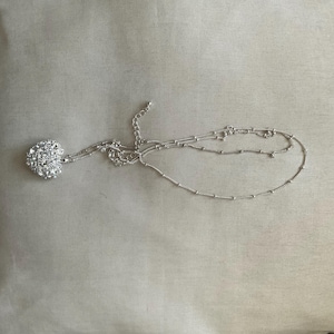 May include: A silver-toned necklace with a heart-shaped pendant covered in small, sparkling rhinestones. The chain is delicate, with small beaded accents and an adjustable clasp. The necklace lies on a neutral-toned surface.