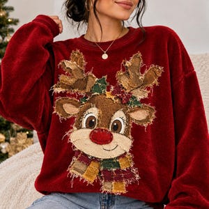 May include: A deep red long-sleeve sweatshirt featuring a large, textured reindeer appliqué on the front. The reindeer has frayed fabric edges, a bright red nose, and a colorful plaid scarf. The design includes green and red fabric accents.