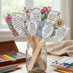 May include: A glass jar containing wooden craft sticks, confetti, and decorative cards with floral designs and Bible verses. The cards feature flower illustrations and inspirational quotes. A wooden pencil case is in the background.