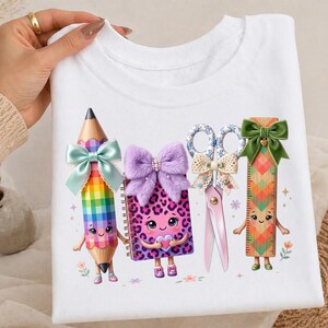 Könnte beinhalten: Ein weißes T-Shirt mit einem Grafikdruck von vier anthropomorphen Schulsachen: ein karierter Bleistift mit hellblauer Schleife, ein lila Notizbuch mit Leopardenmuster und flauschiger lila Schleife, eine Schere mit Spitzenschleife und ein Lineal mit grüner Schleife.