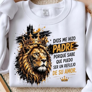 Könnte beinhalten: Ein weißes Sweatshirt mit Rundhalsausschnitt und einer Grafik eines Löwen mit goldener Krone vor einem schwarzen Kreuz. Der Text lautet: DIOS ME HIZO PADRE PORQUE SABE QUE PUEDO SER UN REFLEJO DE SU AMOR. Unter dem Text befindet sich eine kleine goldene Krone.