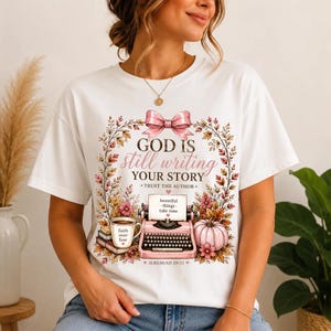 May include: A white t-shirt featuring a graphic print of a pink typewriter, a coffee mug, and a pumpkin surrounded by autumn leaves. The text reads: GOD IS still writing YOUR STORY, TRUST THE AUTHOR, beautiful things take time, and JEREMIAH 29:11.
