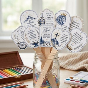 May include: A selection of inspirational cards with religious quotes, presented in a clear glass jar with wooden sticks. The cards feature illustrations of mountains, lions, crosses, and other symbols, with text in a decorative font. A wooden box of coloured pencils and watercolour paints are in the background.