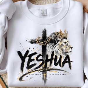 May include: A white crewneck sweatshirt featuring a graphic print of a lion wearing a gold crown next to a black cross with a crown of thorns. The text YESHUA is printed in large black brushstroke letters with SALVATION IS IN HIS NAME below.