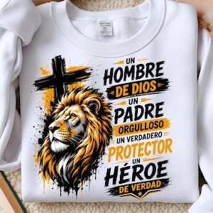 May include: A white crewneck sweatshirt featuring a graphic print of a lion's head and a black cross. The text reads: UN HOMBRE DE DIOS UN PADRE ORGULLOSO UN VERDADERO PROTECTOR UN HÉROE DE VERDAD. The design uses black and yellow ink accents.