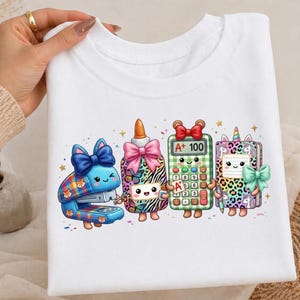 May include: A white t-shirt featuring a colorful graphic of four anthropomorphic school supplies: a blue plaid stapler, a glue bottle with a pink bow, a green checkered calculator displaying A+ 100, and a leopard print notebook with a unicorn.