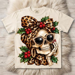 Può includere: Una t-shirt color crema con la grafica di un teschio che indossa un foulard leopardato, occhiali da sole con strass e orecchini a cerchio. Il teschio è circondato da foglie di agrifoglio verdi e bacche rosse, con una mano scheletrica vicino al mento.