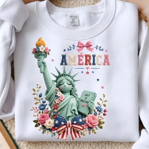 May include: White sweatshirt with a crochet Statue of Liberty design. The design includes the word "AMERICA" and the text "JULY IV MDCCLXXVI". The Statue of Liberty is surrounded by flowers and bows.