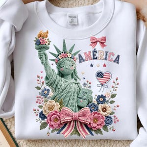 May include: White sweatshirt with a crochet Statue of Liberty design, floral details, a pink bow, and the word "AMERICA". The design includes a heart with the American flag and the text "JULY IV MDCCLXXVI".