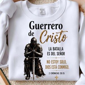 Könnte beinhalten: Ein weißes Langarm-Sweatshirt mit einer Grafik eines knienden Ritters in Rüstung, der ein Schwert hält. Der Text lautet "Guerrero de Cristo", "LA BATALLA ES DEL SEÑOR", "NO ESTOY SOLO, DIOS ESTÁ CONMIGO." und "2 CRÓNICAS 20:15" mit einem kleinen Kreuz.