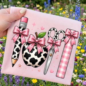 May include: A light pink fabric surface featuring a graphic print of a pencil, an apple, scissors, and a ruler. Each item is decorated with a pink bow. The pencil and apple have a black and white cow print pattern, while the ruler has a pink gingham pattern.