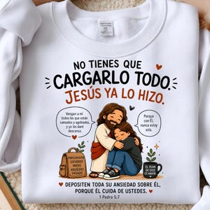 Puede incluir: Una sudadera blanca de cuello redondo con una ilustración gráfica de Jesús abrazando a una persona. El texto dice: NO TIENES QUE CARGARLO TODO. JESÚS YA LO HIZO. Incluye globos de texto en español, una mochila con palabras y una taza.
