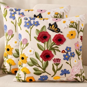 May include: Two square throw pillows featuring an off-white base with colorful floral embroidery. The design includes red poppies, blue forget-me-nots, yellow sunflowers, white daisies, and a black and yellow butterfly scattered across the fabric.