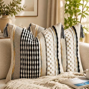 May include: Two square throw pillows featuring a textured beige fabric with vertical stripes in black, white, and yellow. The design includes raised tufted patterns, woven sections, and decorative black and white tassels attached to the top corners.