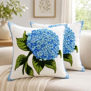 Puede incluir: Dos cojines cuadrados blancos con un diseño de flor de hortensia azul en relieve y hojas verdes. Los bordes de los cojines están rematados con un ribete decorativo de color azul claro, creando un aspecto coherente para la decoración del hogar.