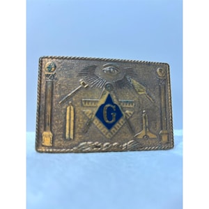 May include: A rectangular brass belt buckle featuring a raised relief design of Masonic symbols, including a central square and compass with a blue enamel letter G, an all-seeing eye, pillars, a gavel, and a trowel, all framed by a rope-style border.