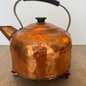 May include: A hammered copper tea kettle with a rounded body, a short spout, and a dark brown handle. The kettle features a matching dark brown knob on the lid and rests on small, dark red feet. The metal surface shows a reflective, textured finish.