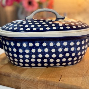 May include: A blue and white polka dot ceramic casserole dish with a lid and handles. The dish is sitting on a wooden cutting board. The lid has a gray handle.