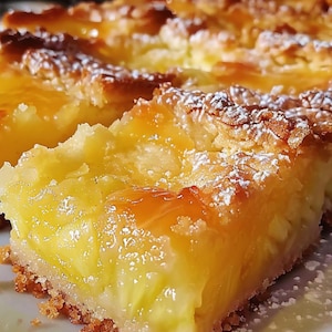 May include: Close-up of a slice of lemon bar dessert. The dessert has a golden-yellow filling with a crumbly crust. The top is dusted with powdered sugar, and the glaze is shiny. The dessert is cut into squares.