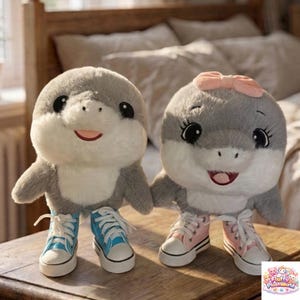 Puede incluir: Dos juguetes de peluche de delfín, uno con un lazo rosa, ambos con zapatillas deportivas. Los delfines son grises con vientres blancos y caras sonrientes. Uno lleva zapatillas azules, el otro rosas. Los juguetes están sobre una superficie de madera.