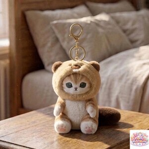 May include: A plush cat keychain wearing a brown bear hood. The cat has large, expressive eyes and a small pink nose. The keychain is made of soft, plush material and has a gold-colored metal ring for attaching to keys or bags.