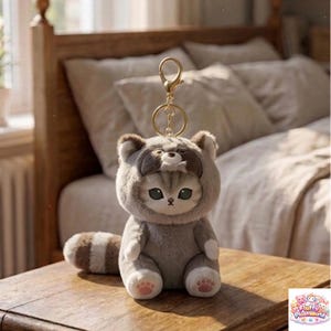 May include: A plush keychain featuring a gray cat wearing a raccoon costume. The cat has green eyes and pink paw pads. The keychain has a gold-colored ring and clasp. The plush toy is sitting on a wooden surface.