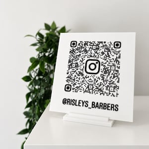 May include: A square white sign featuring a raised black QR code with a central Instagram logo icon. Below the QR code, the text @RISLEYS_BARBERS is displayed in bold black lettering. The sign is supported by a white rectangular base stand.