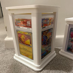 Op de afbeelding: Witte, rechthoekige display case voor verzamelkaarten. De case bevat beoordeelde kaarten, waaronder een Pikachu-kaart met een Gem Mint 10-kwalificatie. De case is ontworpen om trading cards te presenteren en te beschermen.