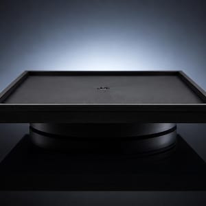 May include: A black, square display stand with a raised, framed top. The stand has a circular base and a dark, matte finish. The background is a gradient of dark gray to light blue.