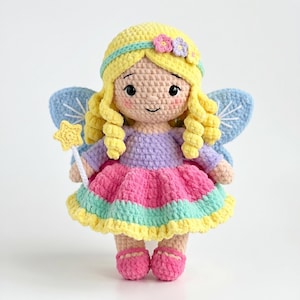 May include: A crocheted doll with yellow hair, a lavender top, and a pink, teal, and yellow skirt. The doll has blue wings, pink shoes, and holds a yellow star wand. It features small pink cheeks, black eyes, and two small flowers on its hair.