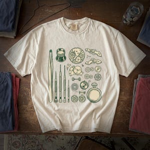May include: A cream-colored short-sleeve t-shirt featuring a dark green graphic print of various watchmaking tools, including tweezers, screwdrivers, a loupe, gears, springs, and a pocket watch case, arranged in a scattered pattern on the front.