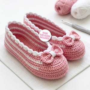 Puede incluir: Un par de zapatillas de ganchillo hechas a mano en un tono rosa suave, con rayas horizontales blancas y un borde festoneado blanco a lo largo de la abertura. Cada zapatilla está adornada con un pequeño lazo rosa en la punta y una etiqueta redonda que dice SHOE SIZE GUIDE INCLUDED.
