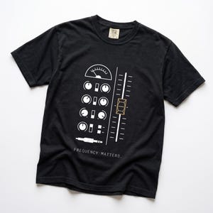 Puede incluir: Una camiseta negra de manga corta con un gráfico blanco de una consola de mezcla de audio con perillas, un fader y un conector jack. El diseño incluye un botón de fader de color dorado y el texto "FREQUENCY MATTERS." impreso en blanco.