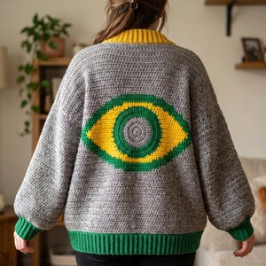 May include: A gray crochet cardigan featuring a large, centered eye motif on the back. The eye design is composed of concentric circles in green, yellow, and gray. The cardigan has a yellow ribbed collar and green ribbed cuffs and hem.