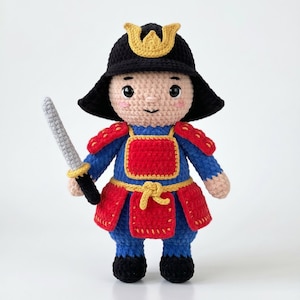 Puede incluir: Una muñeca samurái de ganchillo con un casco negro con cresta dorada, una armadura azul y roja con ribetes amarillos y una espada plateada en una mano. La muñeca tiene una cara de color beige claro con ojos negros y mejillas rosadas.