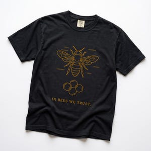 Könnte beinhalten: Ein schwarzes T-Shirt mit kurzen Ärmeln und einer goldenen Strichzeichnung einer Biene, deren Körperteile wie Kopf, Thorax, Abdomen, Antennen, Flügel und Beine beschriftet sind. Darunter befindet sich ein Wabenmuster und der Text IN BEES WE TRUST.