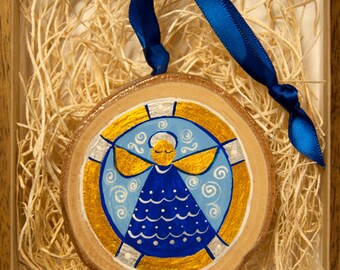May include: A wooden ornament with a blue and gold painted angel design. The angel has a halo and is surrounded by swirls and dots. The ornament is hanging from a blue ribbon.
