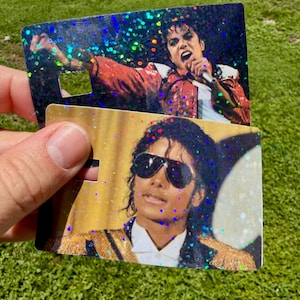 Peut inclure: Deux cartes rectangulaires en plastique avec des effets de paillettes holographiques. La carte du haut montre un artiste en veste rouge chantant dans un micro. La carte du bas affiche un artiste portant des lunettes de soleil et une veste dor&eacute;e.