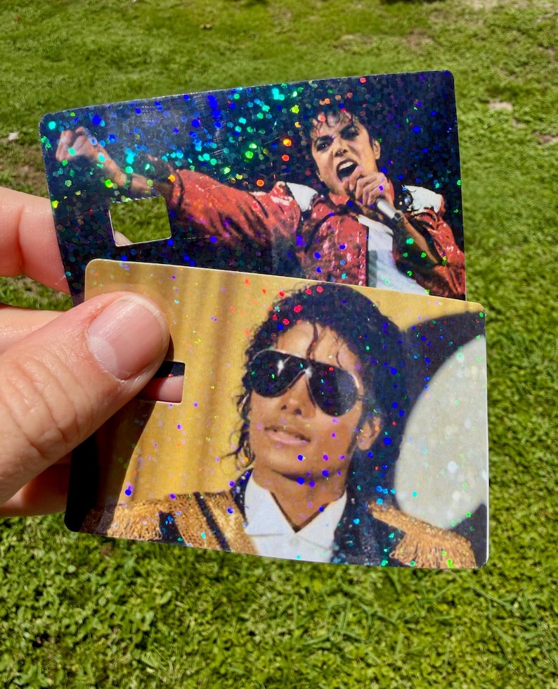 Peut inclure: Deux cartes rectangulaires en plastique avec des effets de paillettes holographiques. La carte du haut montre un artiste en veste rouge chantant dans un micro. La carte du bas affiche un artiste portant des lunettes de soleil et une veste dor&eacute;e.